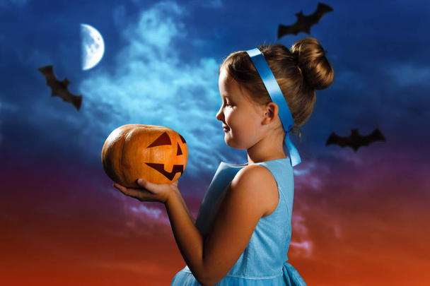 Halloween celebration. Charming little girl in a costume of the Cinderella princess holds a pumpkin on the background of the evening moon sky. - Foto, afbeelding