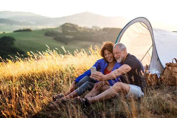 Senior tourist couple with smartphone sitting in nature at sunset, taking selfie. - Foto, Imagen