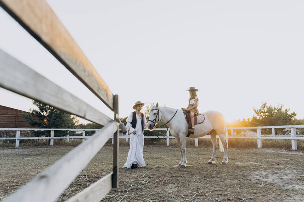 cheerful daughter riding horse while mother holding bridle and walking near on the ranch at the sunset  - Foto, Imagen