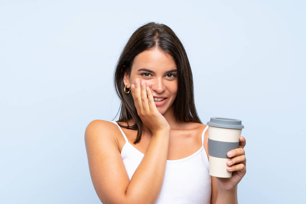 驚きの表情で孤立した青い背景の上にテイクアウトコーヒーを持つ若い女性 - 写真・画像