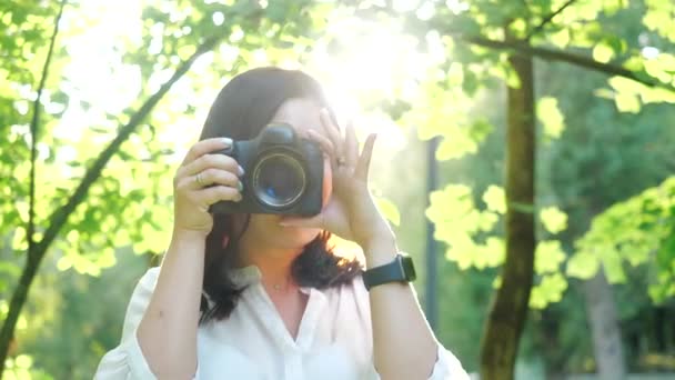 Pretty glimlachend en lachende meisje fotograaf het dragen van witte shirt is het maken van Foto's in een park op een zachte achtergrond van groen gebladerte en sproeiwater. - Video