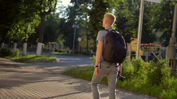 Visão traseira do fotógrafo estudante com uma mochila preta está andando ao longo de uma rua da cidade
 - Filmagem, Vídeo