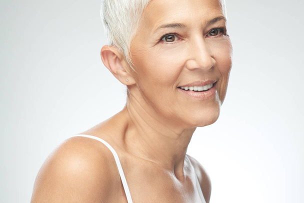 Beautiful smiling senior woman with short gray hair posing in front of gray background. Beauty photography. - Foto, Bild