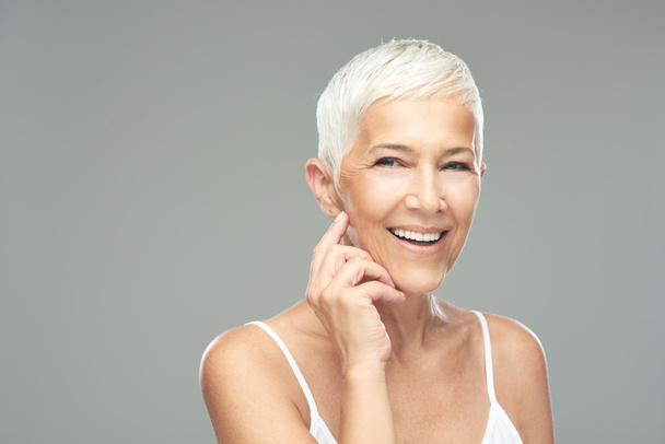 Beautiful smiling senior woman with short gray hair posing in front of gray background. Beauty photography. - Photo, Image