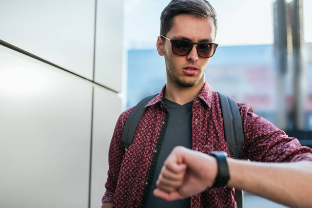 Handsome caucasian man with fashion sunglasses, looking at the clock device on the street. Attractive businessman and tourist walking outdoors in town with backpack. Urban lifestyle concept. - Photo, Image