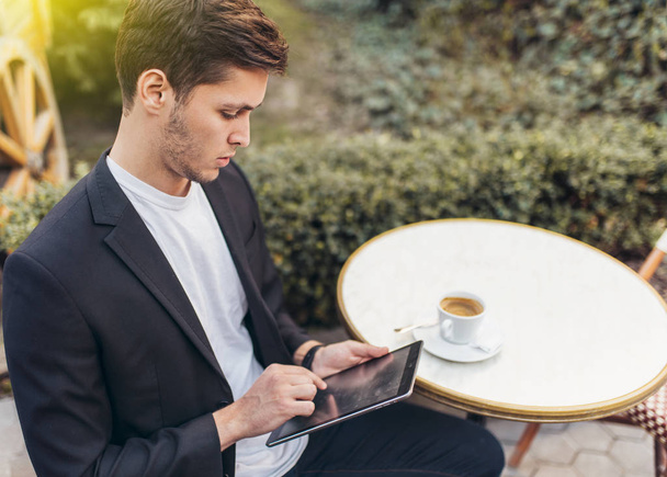Hombre de negocios inteligente usando su tableta mientras desayuna en la cafetería, hombre de negocios sosteniendo una taza de café mientras lee noticias en la tableta por la mañana en la terraza. Concepto de negocio y tecnología
. - Foto, imagen