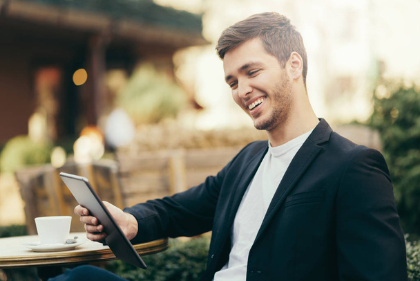 Horizontal, happy caucasian man look to the digital tablet sitting in cafe with cup of coffee. Business man having breakfast sitting on beautiful terrace with plants. Reading and browsing internet. - Foto, imagen
