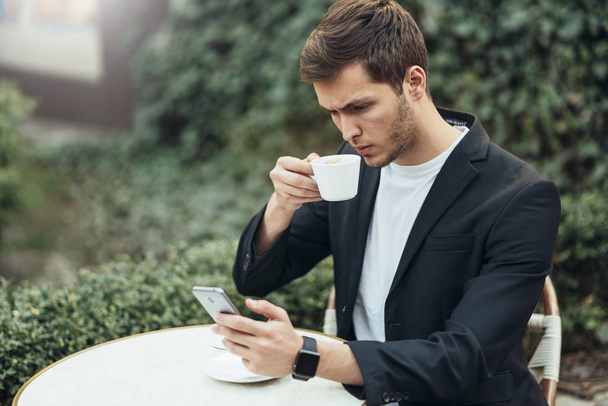 European young and attractive man in casual dress typing messages in social networks.  Business man looks serious, ready to make a sip of fresh brewed coffee at the terrace outdoor in the city. - Photo, Image