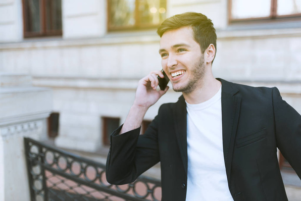 Attractive young business man using mobile phone on building outdoors. Cheerful handsome caucasian man in black suit in the city. Man talking on smart phone on the street. Business lifestyle concept. - Foto, Bild