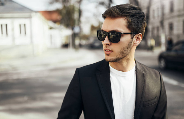 Urban fashion portrait of young handsome businessman in casual suit in dark sunglasses sitting in the street goind to the work and looking aside. - Foto, Bild