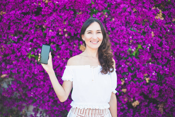 Portrait of happy smiling girl standing on a violet blooming flowers background with smartphone in her hand, romantic summer outfit, outdoors. Happy stunning woman showing her phone. - Фото, зображення
