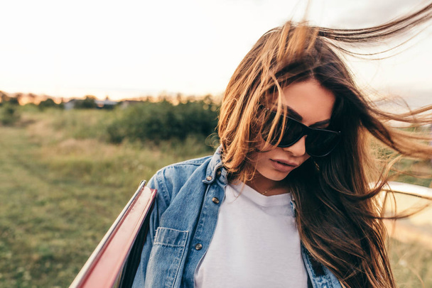 Beautiful attractive young Caucasian woman wearing sunglasses near to her red car on cloudy background. European female outdoor trip with blowing hair on a field background. Travel concept. - Foto, Imagen