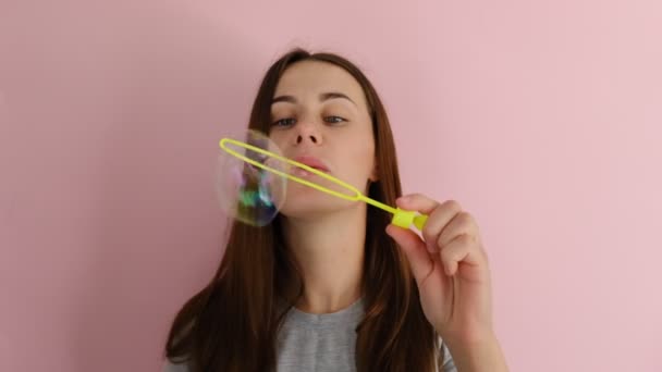 A beautiful cheerful young woman, wearing a t-shirt, standing isolated over pink background, smiling and blowing soap bubbles. People and emotion concept - Video, Çekim