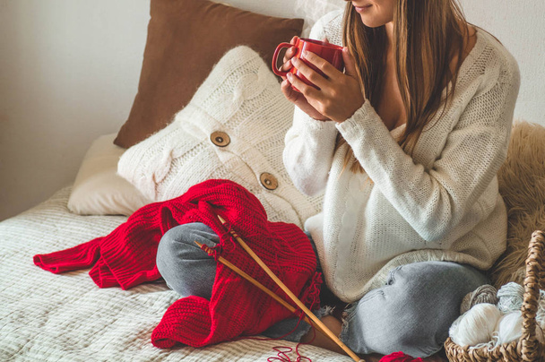 Красивая девушка вяжет теплый свитер с горячей чашкой чая на кровати. Вязание как хобби
 - Фото, изображение
