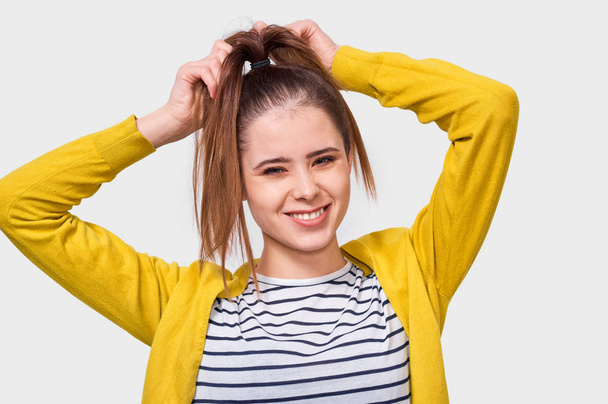 Image of funny young woman collecting hair in a ponytail, wearing casual outfit, posing over white studio wall. Female makes ponytail, advertises healthy natural hair and looks up to copy space. - Foto, Imagen