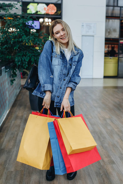 Beautiful young woman in shopping center posing on the background of boutiques. Beauty smiling woman in a denim jacket with shopping bags standing in shopping mall. Shopper, sales, shopping center - Foto, immagini