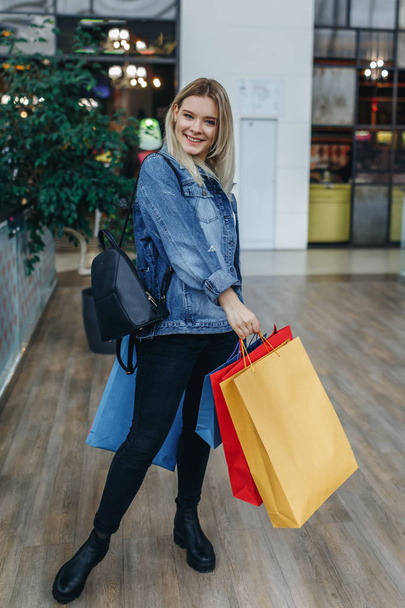 Mode winkelen jonge blonde vrouw portret op de achtergrond van boetieks. Schoonheid lachende vrouw in een denim jasje met boodschappentassen staan in het winkelcentrum. Shopper, verkoop, winkelcentrum - Foto, afbeelding