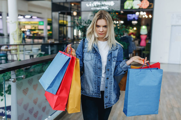 Young serious woman in shopping center posing on the background of boutiques. Beauty woman in a denim jacket with colorful shopping bags in shopping mall. Sale, gifts, holidays and people concept - Photo, Image