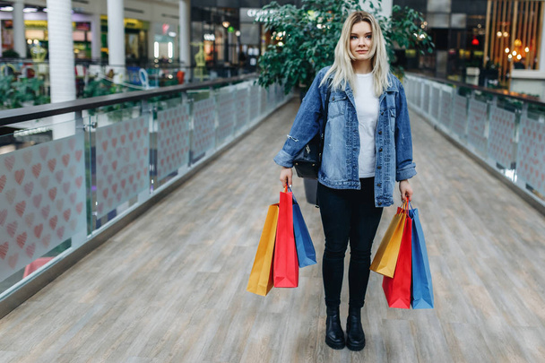 Butiklerin arka planında alışveriş merkezinde genç kadının portresi. Alışveriş merkezinde renkli alışveriş çantaları ile kot ceketli güzellik kadını. Sol taraftaki alanı kopyalama - Fotoğraf, Görsel