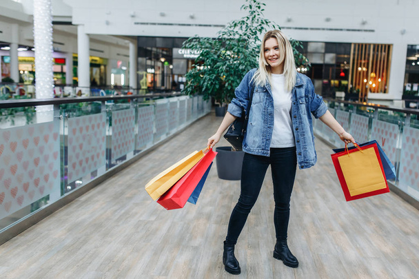 Young woman in shopping center on the background of boutiques and looking aside. Beauty woman in a denim jacket with colorful shopping bags in shopping mall. Copy space on the left side - Photo, Image