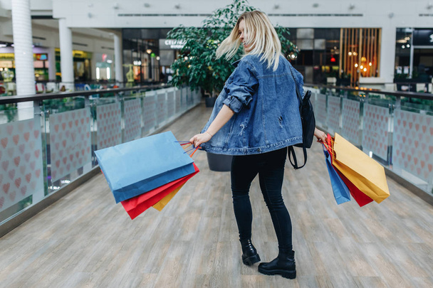 Alışveriş merkezinde kameraya geri yürüyen genç kadın butiklerin arka planında. Alışveriş merkezinde renkli alışveriş çantaları ile kot ceketli güzellik kadını. Sol taraftaki alanı kopyalama - Fotoğraf, Görsel