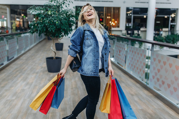 Woman after shopping. Cheerful young woman in shopping center on the background of boutiques. Beauty woman in denim jacket with shopping bags in shopping mall. Copy space on left or right side - Foto, Imagem