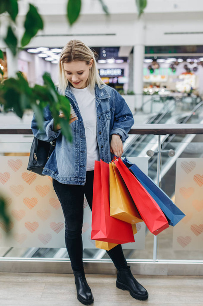 Cep telefonu ile moda alışveriş genç kadın Portresi. Alışveriş merkezinde duran alışveriş çantaları ile kot ceketli güzellik kadını. Alışveriş, satış, alışveriş merkezi - Fotoğraf, Görsel
