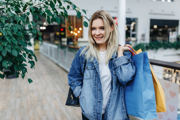 Time for shopping and spending time in trade center. Pretty woman in a denim jacket standing in a shopping mall with coloured shopping bags in hands. Shopping, sale, gifts concept, copy space - Fotó, kép