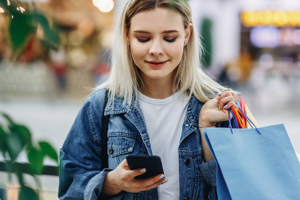 Κοντινό πορτραίτο της όμορφης ξανθιάς νεαρής γυναίκας στο εμπορικό κέντρο χρησιμοποιώντας το κινητό smartphone. Μια όμορφη γυναίκα με ένα τζιν μπουφάν με σακούλες για ψώνια που στέκονται στο εμπορικό κέντρο. Αγοραστής, εμπορικό κέντρο - Φωτογραφία, εικόνα