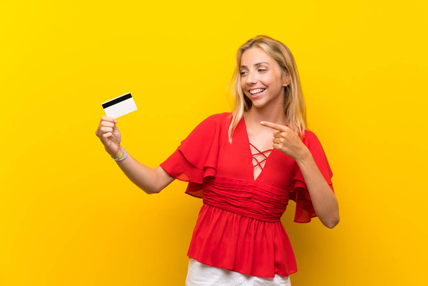 Blonde young woman over isolated yellow background holding a credit card - Photo, image
