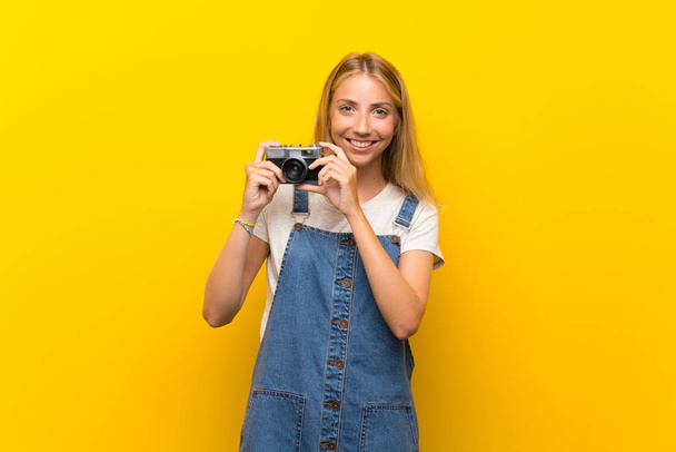 Blonde young woman over isolated yellow background holding a camera - Foto, Bild