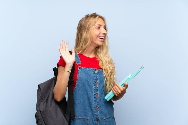 Молода блондинка студентка над ізольованою синьою стіною з несподіваним виразом обличчя
 - Фото, зображення