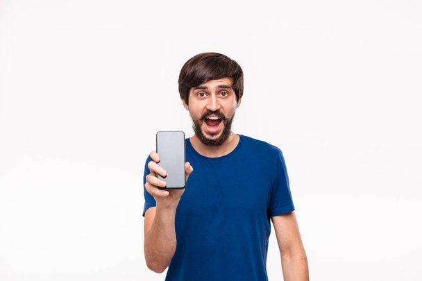 白い背景の上に隔離されたカメラでスマートフォンの画面を示すひげと口ひげを持つ青いシャツを着た格好良いブルネットの男。驚きの感情とジェスチャー. - 写真・画像