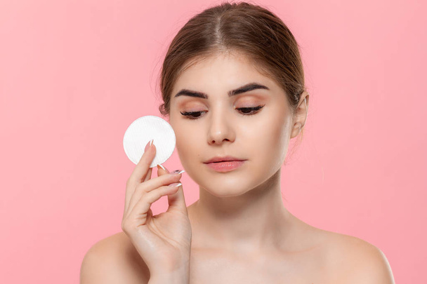 Beautiful model applyes cosmetic product to skin with white cotton sponge isolated over pink background. Concept of beauty and health treatment. - Foto, imagen