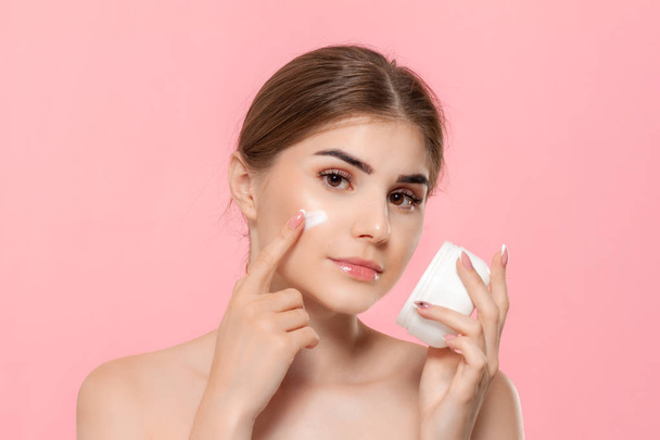 Beautiful girl applying moisturizing cream from tube to skin. Model looks at the camera isolated over pink background. Concept of beauty and health treatment. - Foto, imagen