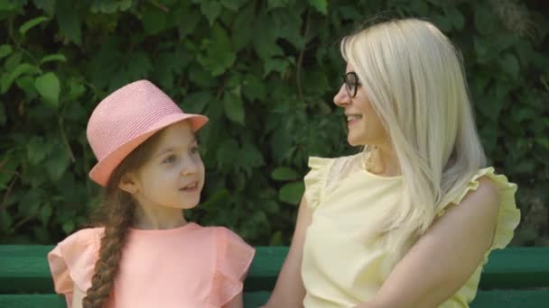 Cute mature blond mother in glasses and her little daughter chatting while sitting on the bench in the summer park. Happy loving family. Woman and girl together outdoors. - Záběry, video