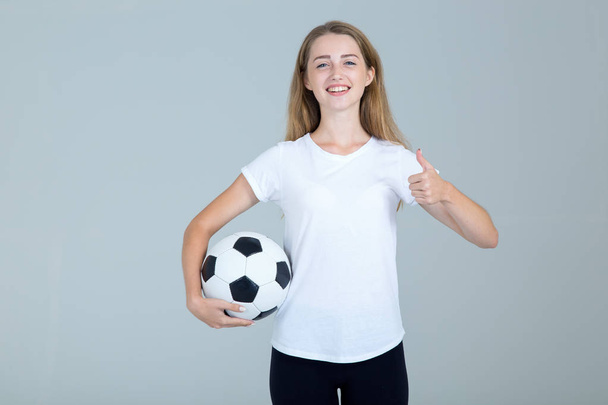 Happy young woman with a soccer ball in her hands shows thumb up standing on a gray background. Football fan. - Fotografie, Obrázek