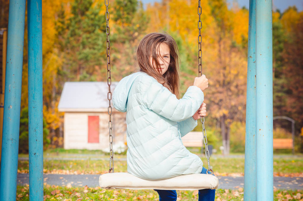 涼しい秋の日に遊び場でブランコに座ってダウンジャケットに身を包んだかわいい女の子. - 写真・画像