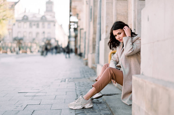 Outdoor portrait of a young beautiful fashionable lady sitting on stairs of old building on a street of the old city. Model wearing stylish clothes. Female fashion concept. City lifestyle. - 写真・画像