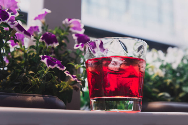 Beautiful red cocktail with ice near the window. Glass standing next to beautiful purple flowers. Relax with negroni. - Photo, Image