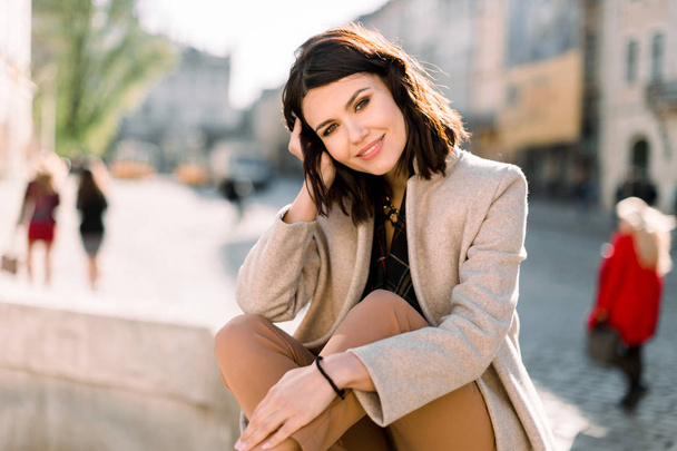 Photo of beautiful stylish brunette woman in the old city, wearing beige coat, brown pants. Fashion summer photo, close up, copy space - Valokuva, kuva