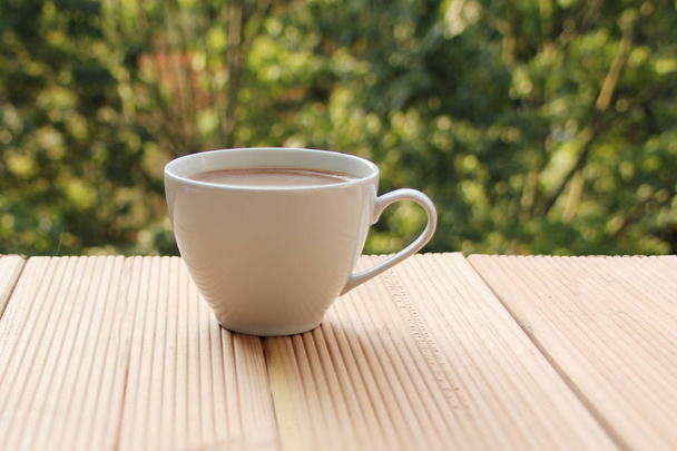 white cup with coffee on a light wooden table against a green summer garden, close-up, copy space - Фото, зображення