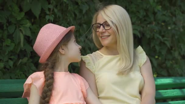 Mature blond mother in glasses and her little daughter looking at camera while sitting on the bench in the summer park. Happy family. Woman and girl together outdoors. - Video