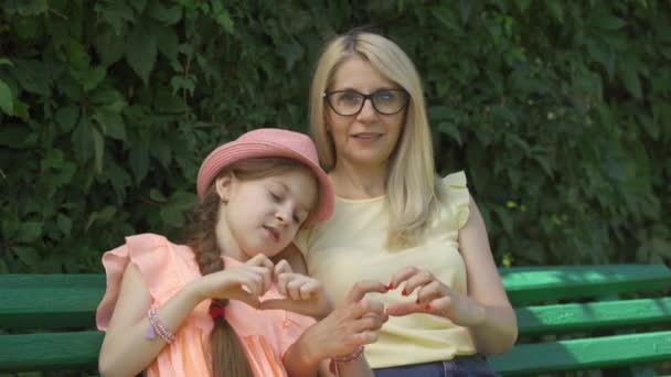Mature blond mother in glasses and her daughter forming heart shape with their fingers looking at camera sitting on the bench in the summer park. Happy family. Woman and girl together outdoors. - Кадры, видео