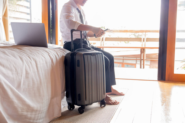 Businessman holding cellphone while sitting on bed with suitcase in hotel room during business trip. - Photo, image