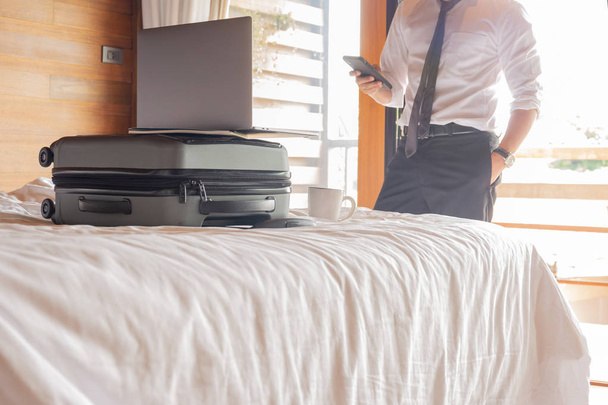 Businessman looking at cell phone with laptop on top of suitcase on the bed. - Photo, Image