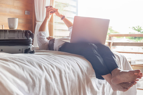 Расслабленный бизнесмен лежит в постели и смотрит на телефон с ноутбуком на коленях.
. - Фото, изображение
