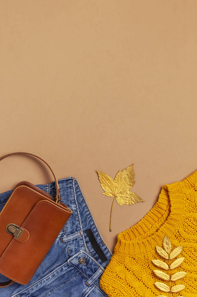 herbstliches Modekonzept. braune Damentasche aus Leder, orangefarbener Strickpullover, blaue Jeans, goldenes Herbstblatt auf braunem Hintergrund von oben. modische Damenaccessoires Damenkleidung - Foto, Bild