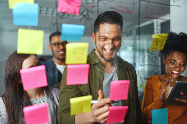 Group of smiling casual business people planning and writing project and ideas over the sticky notes pasted on glass wall at modern office - Photo, image