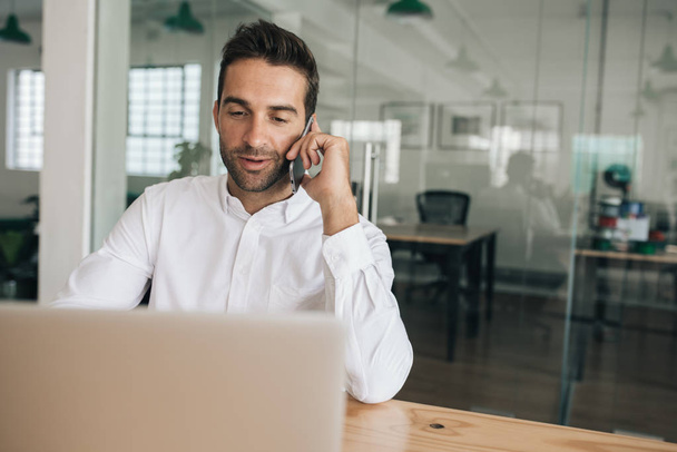 Улыбающийся молодой бизнесмен разговаривает с клиентом по мобильному телефону, сидя за столом в современном офисе, работая над ноутбуком
 - Фото, изображение